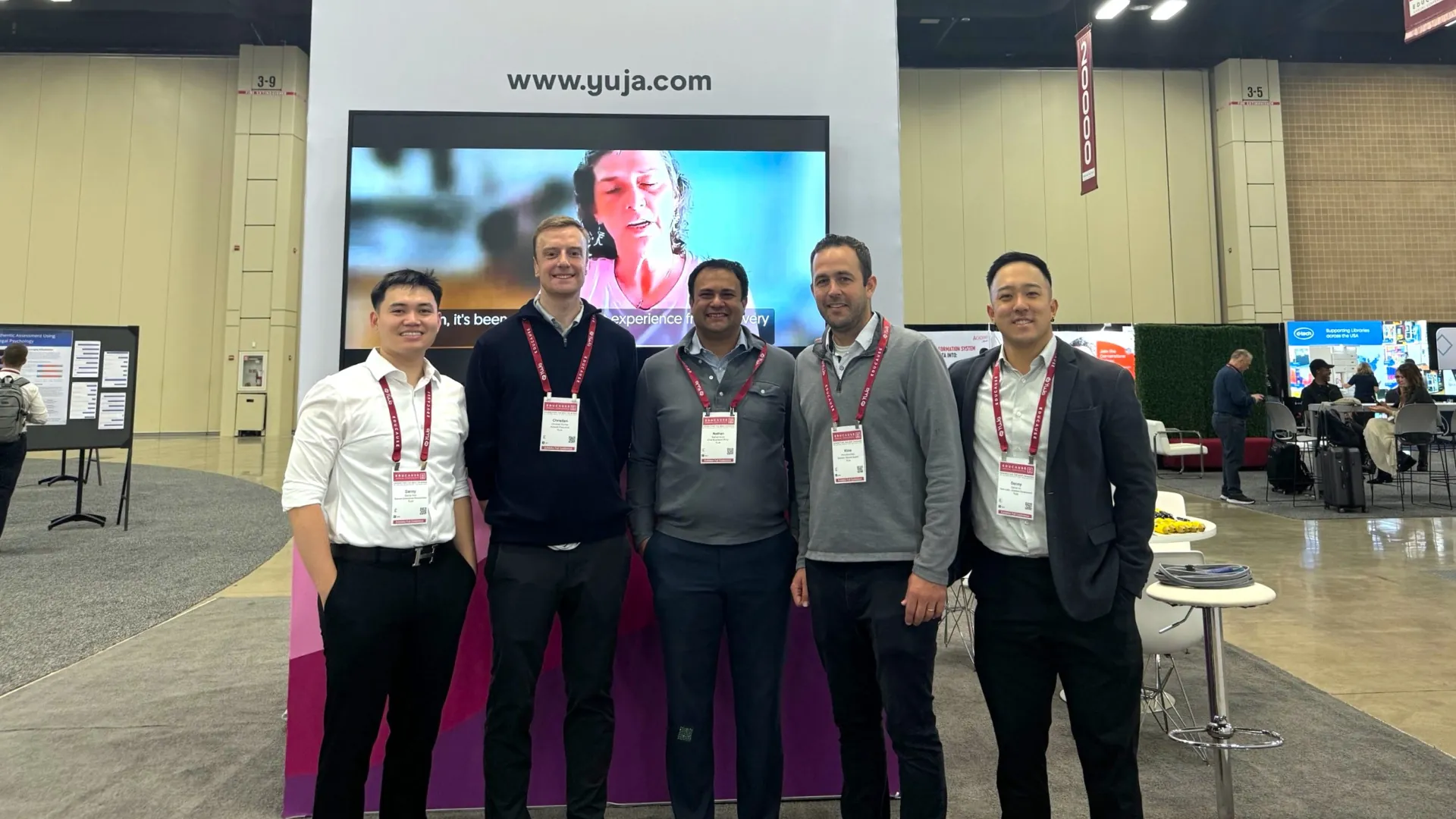 YuJa team members standing in front of their booth, featuring a banner with the website URL www.yuja.com.
